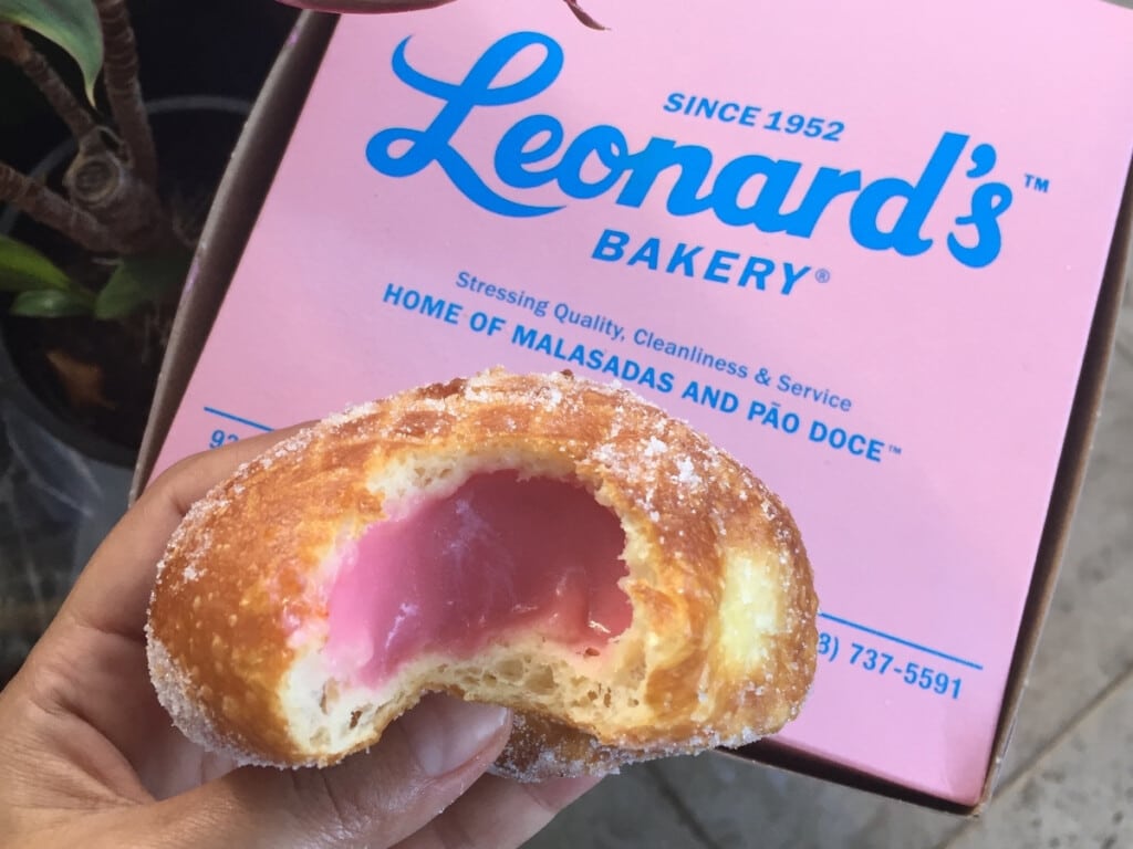 Malasada with pink filling in front of pink box of Leonard's Bakery Malasadas.