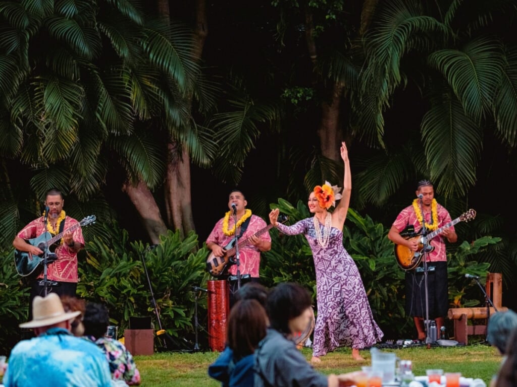 Pre-show performance at Waikiki Starlight Luau: Ocean Lawn, featuring hula dancer and a Hawaiian band in colorful aloha shirts and lei playing live music surrounded by tropical greenery.