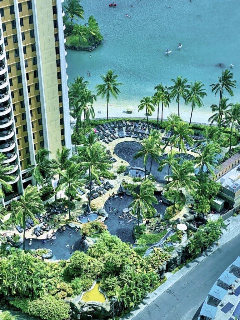 A scenic view of the Hilton Hawaiian Village Paradise Pool in Waikīkī with the Duke Kahanamoku Lagoon in the background, surrounded by swaying palm trees, sun loungers, and lush tropical greenery, with the hotel’s high-rise building on the left overlooking the pool.