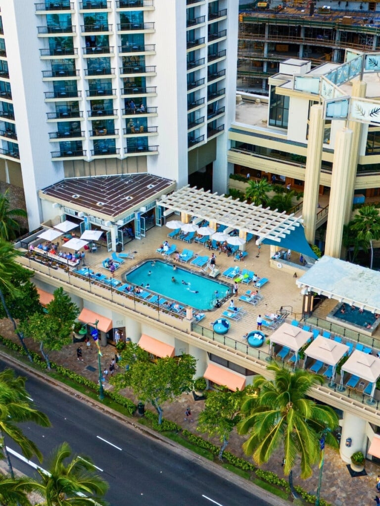 Arial view of the pool area at Hyatt Regency Waikiki Beach Resort and Spa featuring cabanas, a bar, and lounge seating. With Kalākaua Avenue in the foreground and the hotel as a backdrop, the luxurious pool area has aqua colored beach chairs that match the color of the pool.