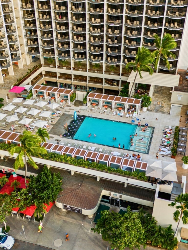 Arial shot of the pool area of Waikiki Beach Marriott Resort & Spa. This is set against the backdrop of the high rise hotel with Cabanas and white beach umbrellas lining the pool area.