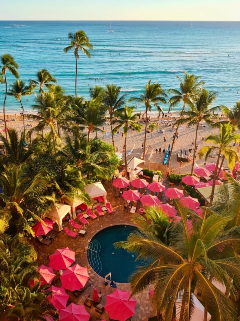 Aerial view of the Malulani Pool at The Royal Hawaiian resort, set against a stunning backdrop of Waikīkī Beach with the blue ocean and palm trees. Pink sun lounges, umbrellas and cabanas line the circular pool.