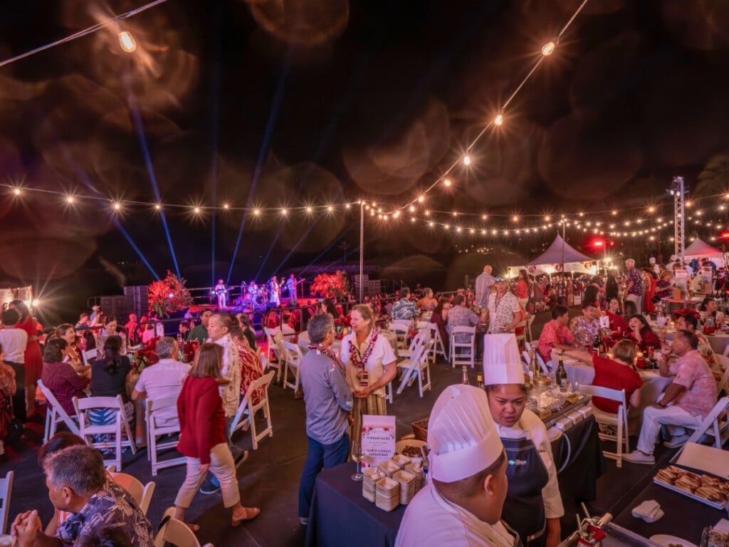 Evening event at the Hawaiʻi Food & Wine Festival. There is chefs in the forrgound and guests seated enjoying food and watching live music.