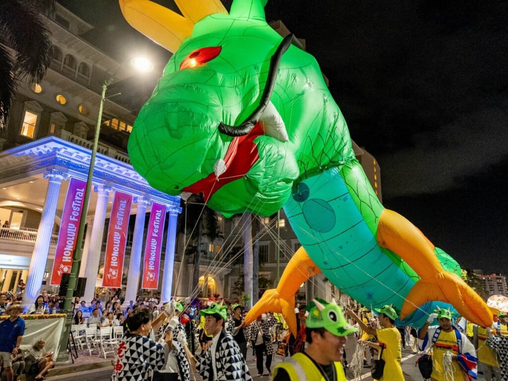 A vibrant dragon float parades down Kalakaua Avenue during the Honolulu Festival and crowds gather beneath brightly lit banners, enjoying the festive atmosphere and performances.
