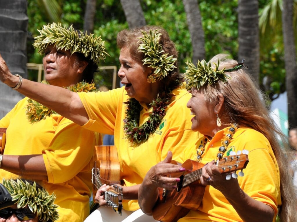 Musicians dressed in yellow and wearing tradition lei at the Prince Kūhiō Celebration parade in Waikīkī. They are smiling and waving while playing ukuleles.
