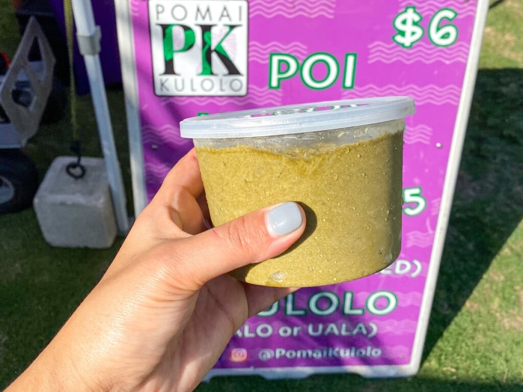 Hand holding tub of poi in front of a purple Pomai PK Kulolo sign showing $6 price at Honolulu Farmers Market.