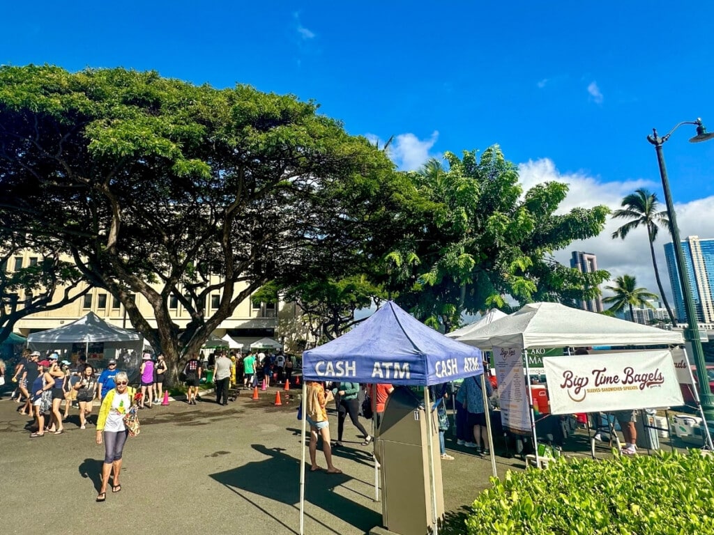 Sunny day at Kaka’ako Farmers Market with stalls under the trees and palm trees.