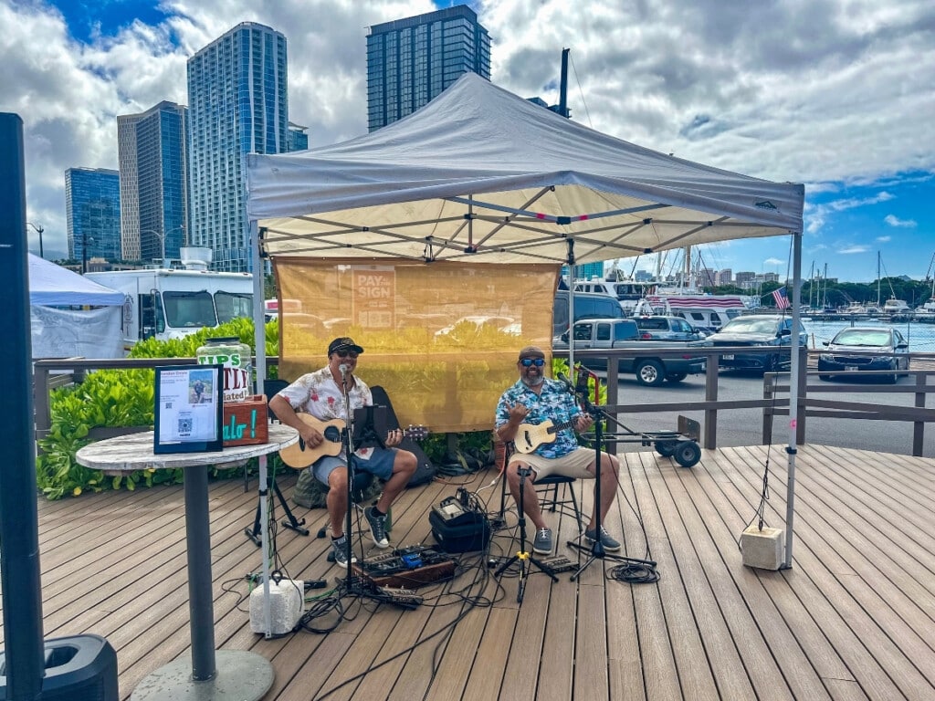 Live music with a guitarist and musician playing the ukulele on a stage by the boat Harbour at Kaka’ako Farmers Market.