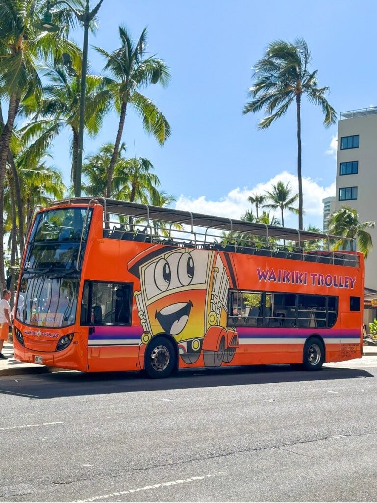 A big orange Waikiki Trolley picking up passengers on Kalākaua Ave. at Duke Kahanamoku Statue in Waikīkī.