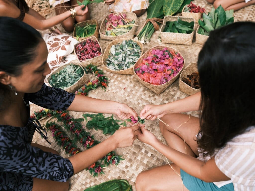 Two participants crafting a beautiful lei together in a lei-making class, surrounded by colorful flowers and bamboo woven containers.