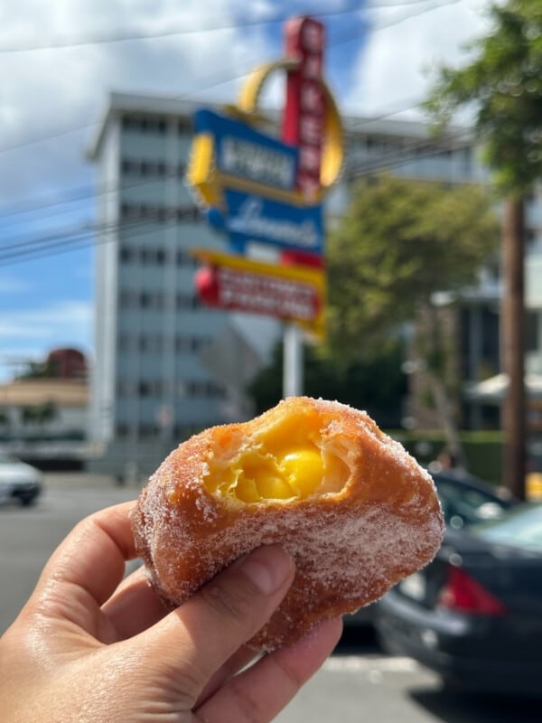 A hand holding a malasada (Portugese doughnut) with a mango (yellow) filling in front of the Leonard's Bakery sign in Kapahulu, O'ahu.