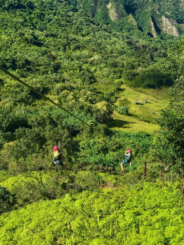 Two people ziplining across tropical greenery at Kualao Ranch.