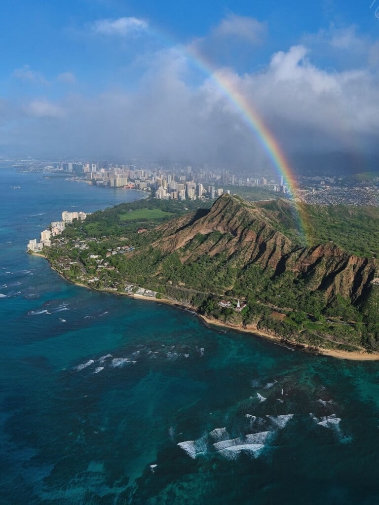 Panoramic aerial view of Diamond Head Crater with rainbow in the foreground and Waikīkī cityscape in the background.