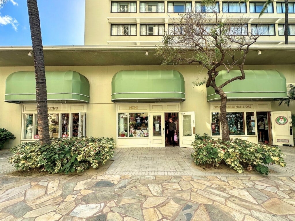 Moana Surfrider shopping area in Waikīkī featuring boutique stores including San Lorenzo Bikinis & Waikiki Christmas Store with green awnings and elegant store fronts.