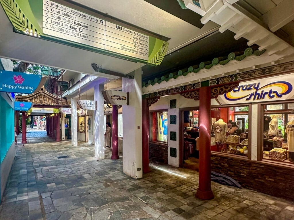Pathway at Hilton Hawaiian Village shopping area in Waikīkī, Hawaiʻi featuring Crazy Shirts and other boutique stores, with tiled stone flooring, traditional architectural accents, and tropical signage.