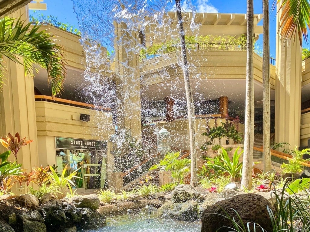 Scenic view of the Pualeilani Atrium Shops at Hyatt Regency Waikiki Beach Resort, featuring a vibrant tropical courtyard with a fountain, lush greenery, and open-air architecture.