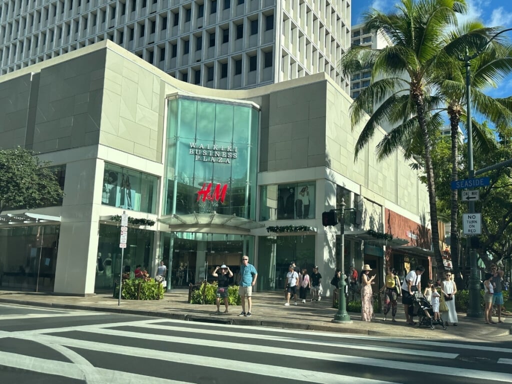 Exterior view of Waikiki Business Plaza a shopping center in Waikīkī  that is multi-story building on a main street with palm trees close by.