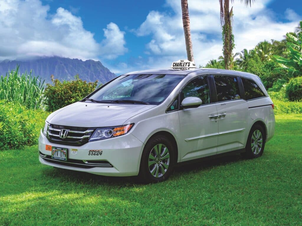 A white Charley's Taxi Honda Odyssey vehicle with a lush Oʻahu botanical garden backdrop.