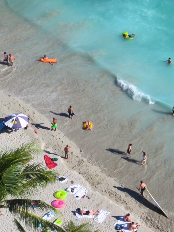 Aerial shot of Waikīkī Beach with palm trees and people sunbaking and floating in the water with colorful floats.
