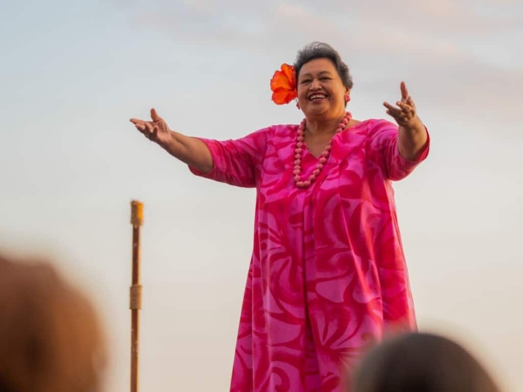 A skilled kumu hula, an elegant woman in her 70s dressed in pink, performs gracefully on stage as the sun sets at the Ahaʻaina Luau at the Royal Hawaiian Resort.