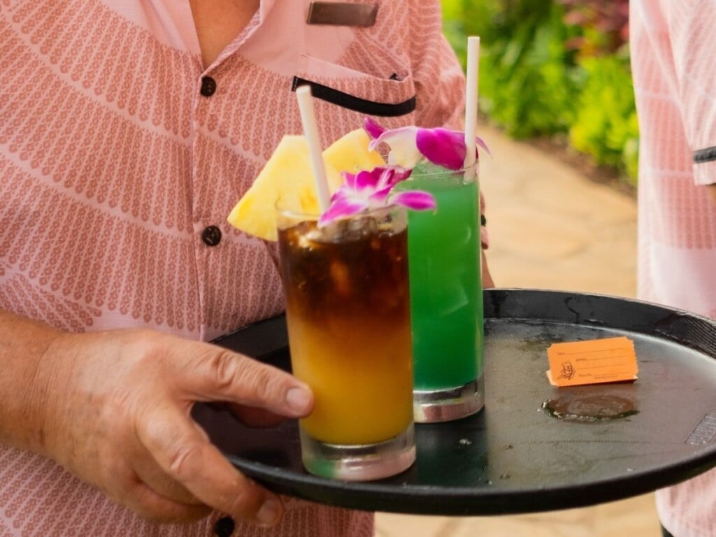 At a ʻAhaʻaina, A Royal Hawaiian Lūʻau, a server brings out two vibrant cocktails, each topped with a delicate purple orchid.