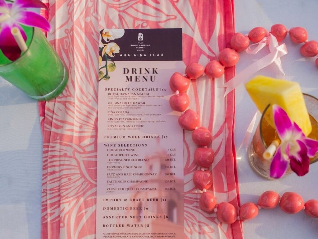 The drink menu rests on a table at the ʻAhaʻaina, A Royal Hawaiian Lūʻau, surrounded by two vibrant cocktails and a kukui nut lei.