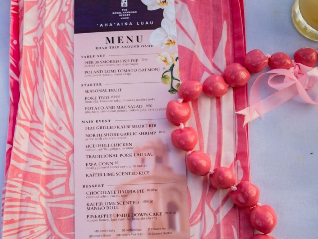 Ahaʻaina Luau at the Royal Hawaiian Resort features a pink menu on a pink tablecloth adorned with Kukui Nut lei.