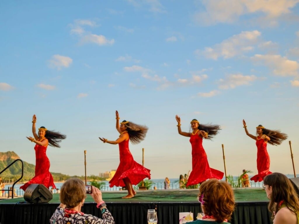 Dancers in red dresses perform hula against the backdrop of Diamond Head as the sun sets during the Ahaʻaina Luau at the Royal Hawaiian Resort.