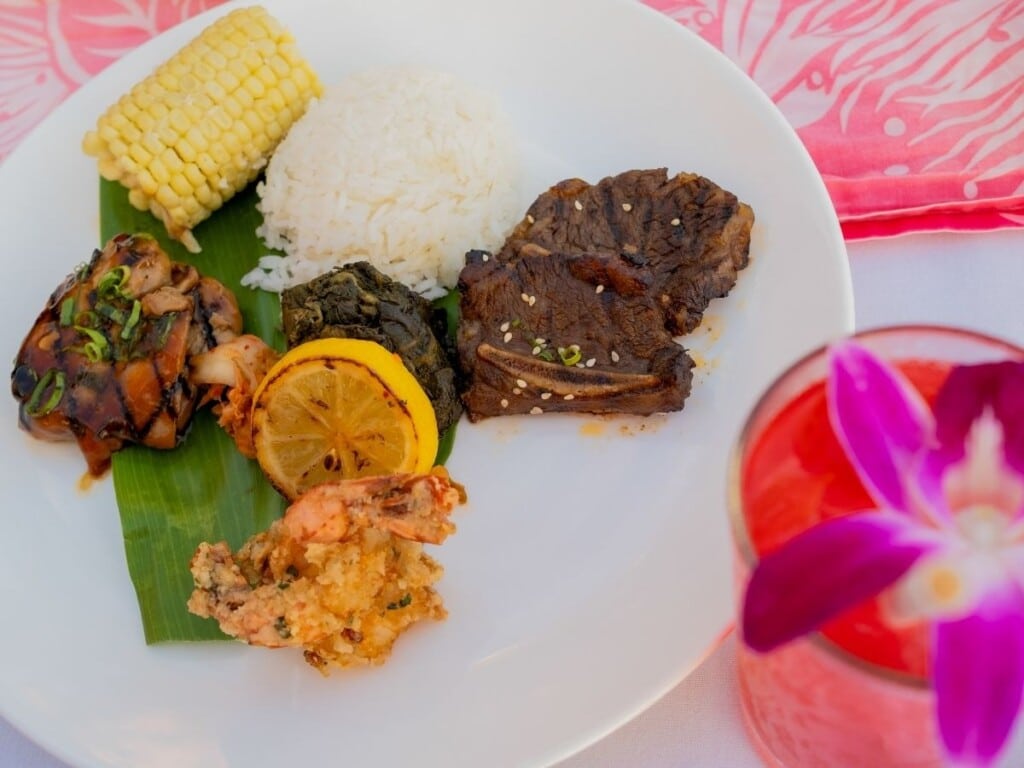 A luau plate with Fire-Grilled Kalbi Short Ribs, North Shore Garlic Shrimp, Huli Huli Chicken, Traditional Pork Lau Lau, Ewa Corn and Rice with a tropical cockatil on the side.