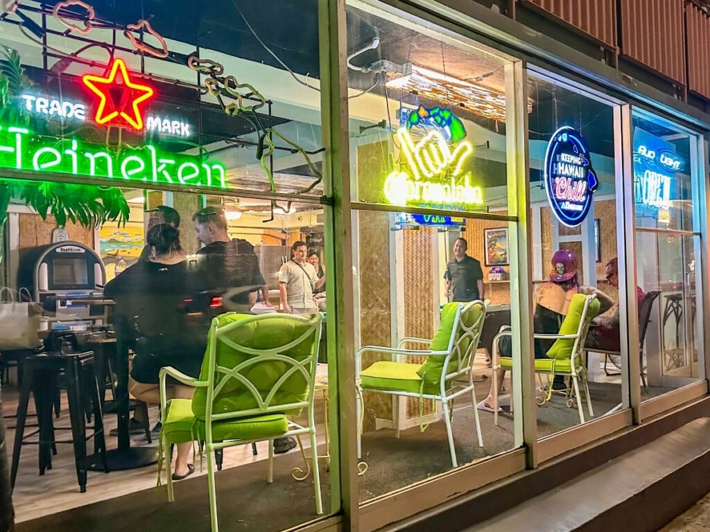 Street view of Honolulu Tavern, a classic dive bar in Waikīkī, featuring neon signs, casual seating, and a laid-back atmosphere.
