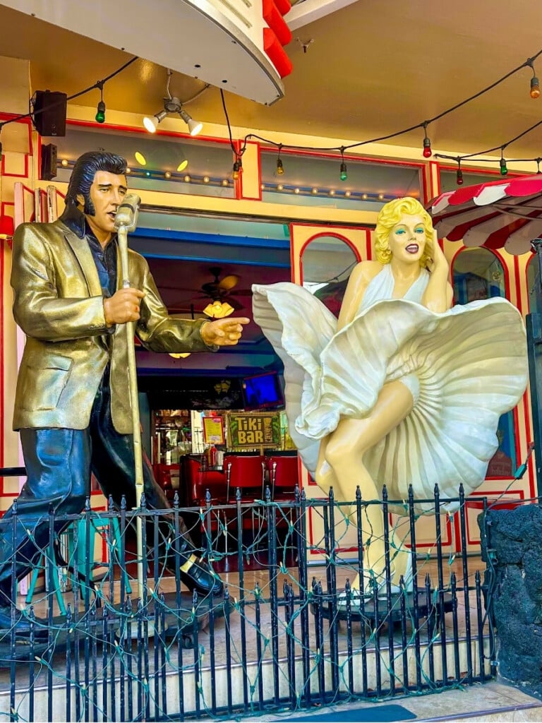 The exterior of Rock Island Café in Waikīkī featuring iconic statues of Elvis Presley and Marilyn Monroe, giving the restaurant a lively, retro Hollywood vibe.