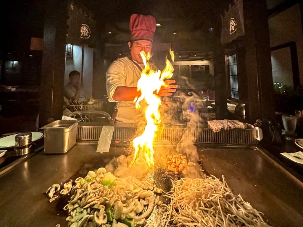 A chef at Tanaka of Tokyo in Waikīkī cooks teppanyaki, with flames lighting the grill. Fresh vegetables, noodles, and seafood sizzle on the hot iron surface, creating an immersive dining experience.