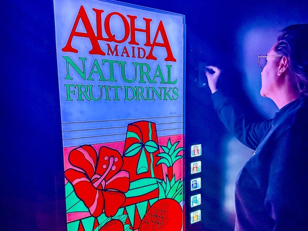 Dimly-lit area leading to the Wild Orange Speakeasy Bar in Waikīkī, featuring an ‘Aloha Made Natural Fruit Drinks’ vending machine. A person is inserting a nickel into the machine to enter the bar.