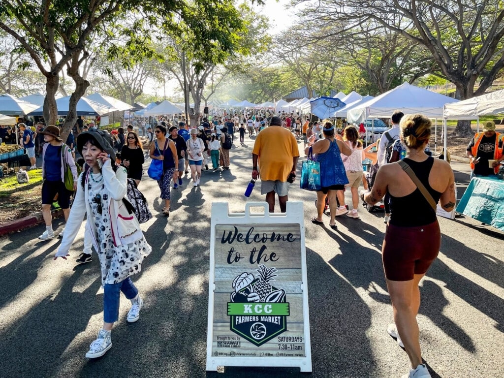 The busy KCC Farmers Markets at Diamond Head with signage reading "Welcome to the KCC Farmers Market).