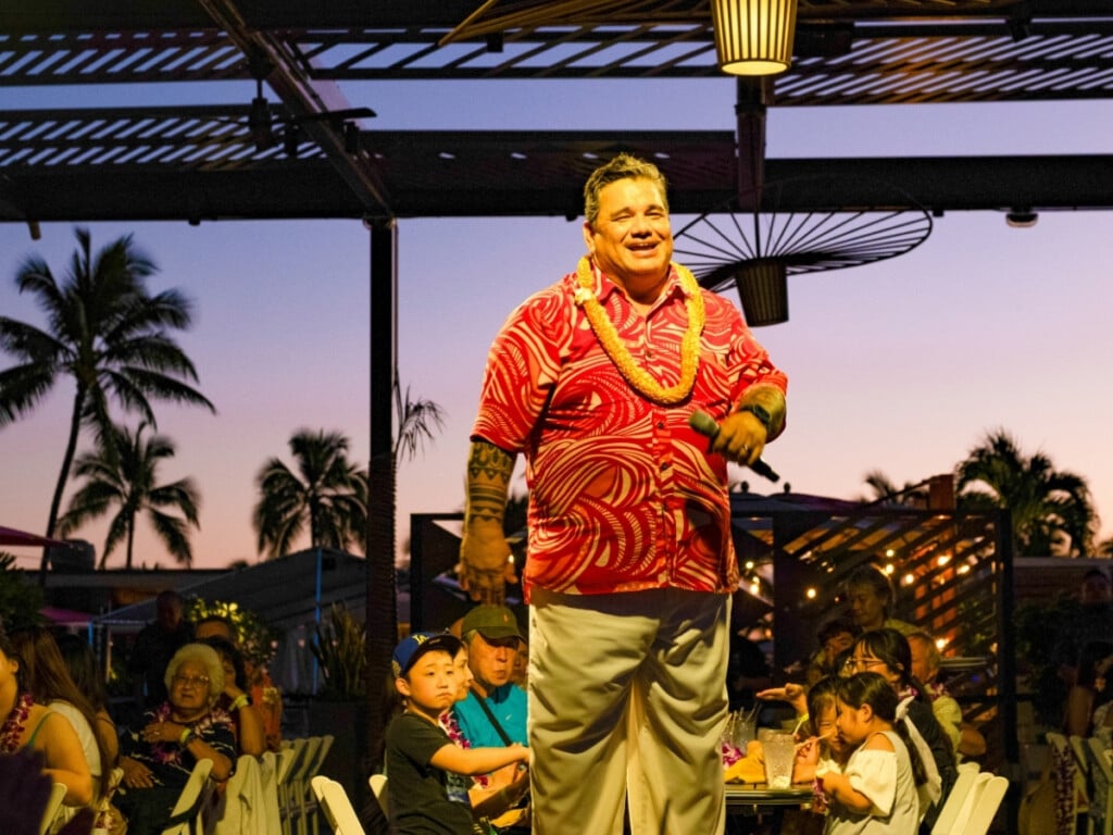 The Master of Ceremonies (MC) on stage at Pāʻina Waikīkī Luau featuring vibrant sunset in the background.