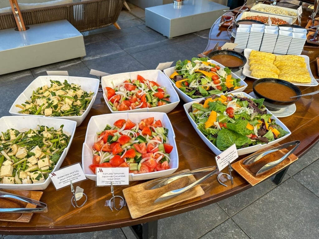 The salad section at at Pāʻina Waikīkī Luau including Watercress and Tofu Maui Onion Salad, Kamuela Tomatoes, Japanese Cucumber & Maui Onion Salad, Fresh Island Greens, Shaved Kabocha, Fennel, Tomatoes & Ume-Lilikoi Vinaigrette and Sliced Fresh Island Maui Gold Pineapple.