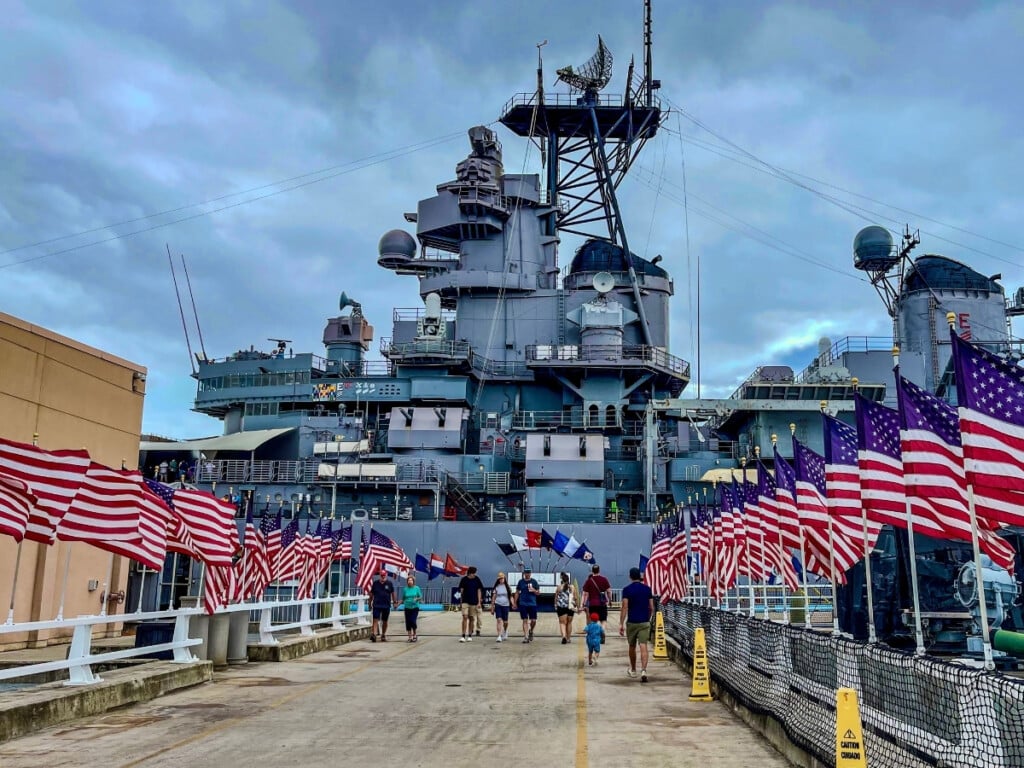 Entrance of the Battleship Missouri Memorial with pathway lined with American flags and the ship in the background at Pearl Harbor.