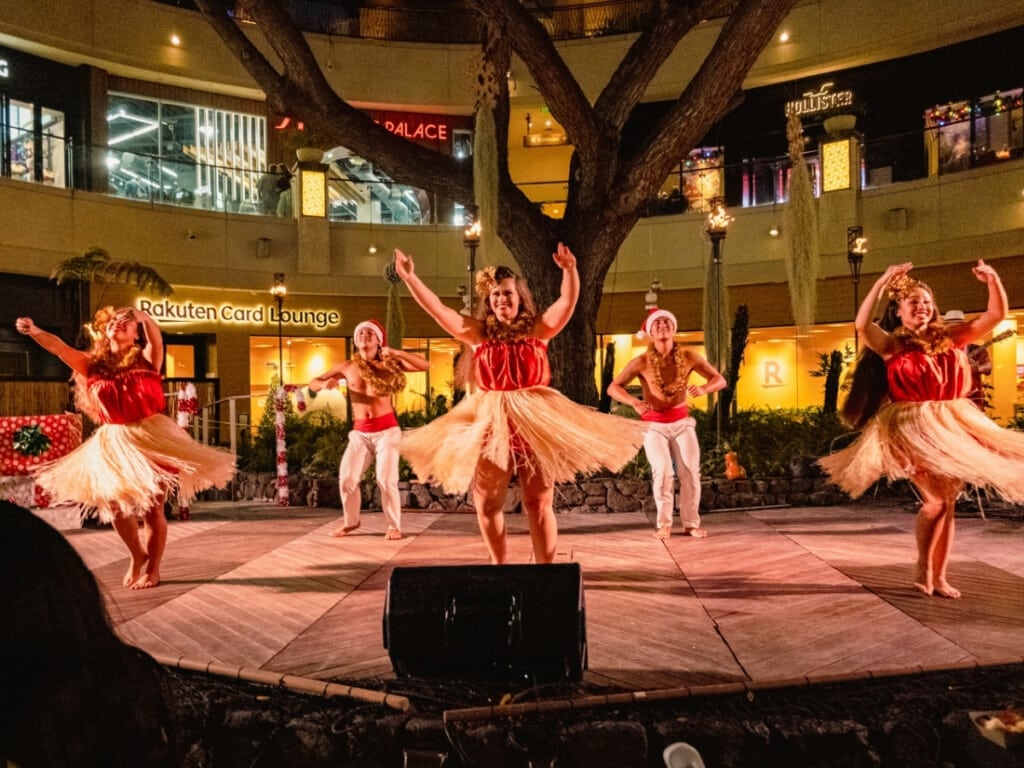 Dancers wearing grass skirts and red and white attire perform underneath the banyan tree at the Queens Waikiki Luau.