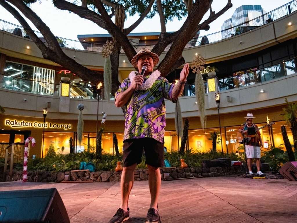 MC welcomes guests featuring banyan tree and guitarist in background at Queens Waikiki Luau.