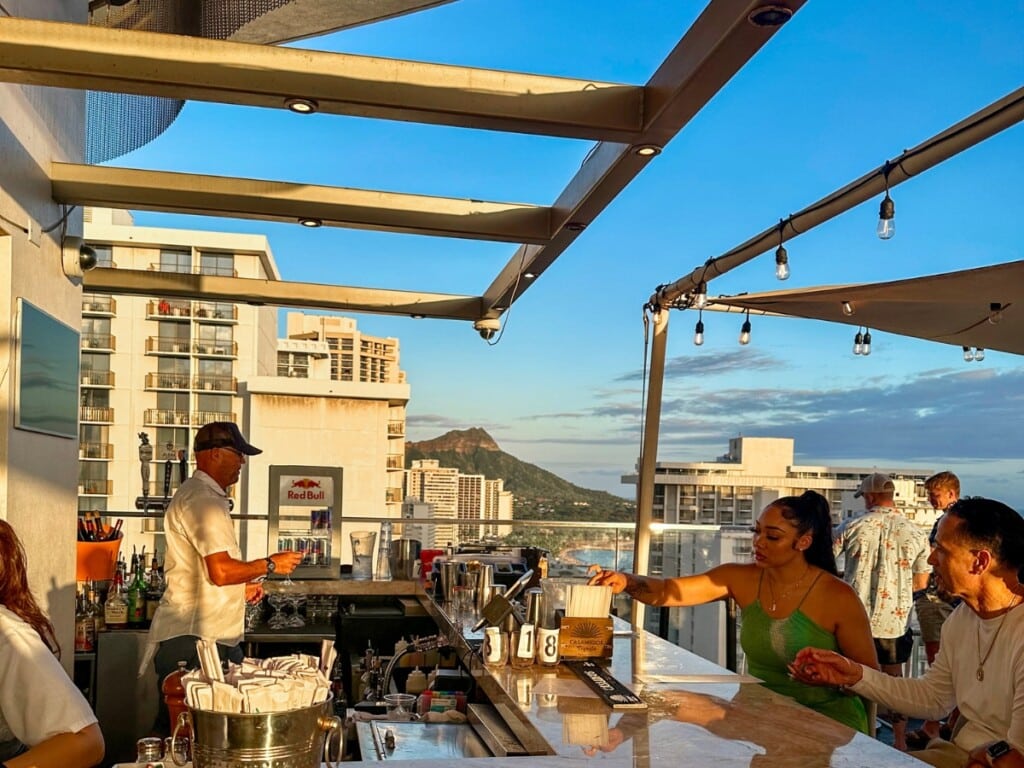 Bar tender serving guests drinks at SKY Waikiki's Bar with a view of diamond head in the background.