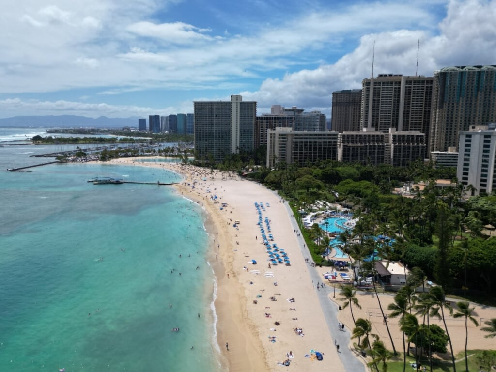Aerial view of Duke Kahanamoku Beach in Waikīkī on a sunny day with palm trees and hotels lining the shoreline.