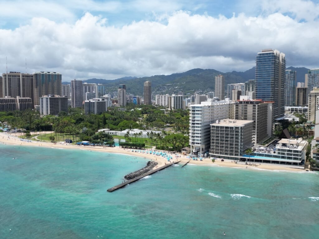 Aerial view of Fort DeRussy Beach Park in Waikīkī with hotel buildings and the mountains in the background.
