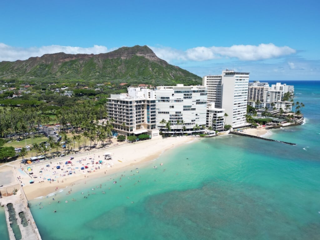 Kaimana Beach in Waikīkī with swimmers and surfers enjoying the clear blue water, framed by tall palm trees. White hotel buildings line one corner of the shore, with the iconic Diamond Head crater towering in the background.
