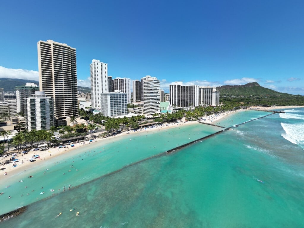 Aerial view of Kuhio Beach Park in Waikīkī, with surfers in the water, palm trees along the shoreline, and the towering Diamond Head crater in the background.