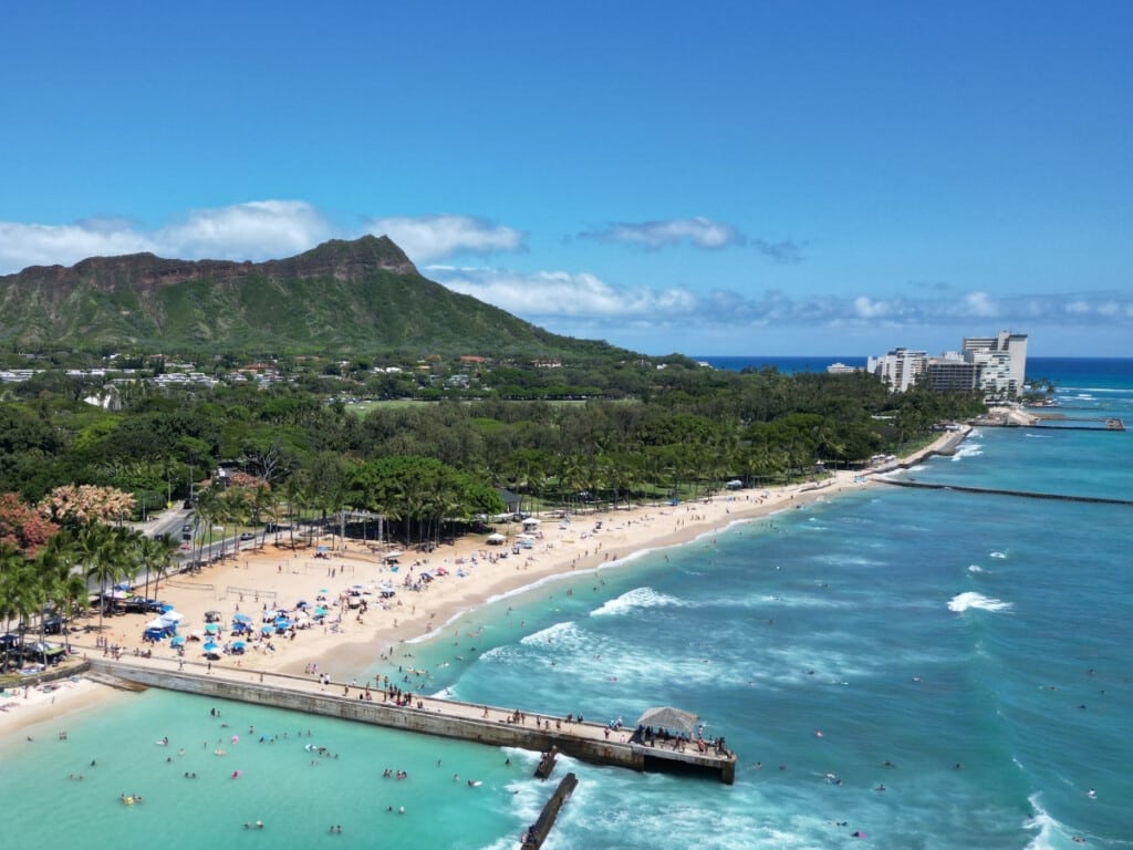 A sunny day at Queens Beach in Waikīkī, with bright blue beach umbrellas dotting the golden sand. Palm trees and lush greenery frame the beach, while Diamond Head rises majestically in the background.