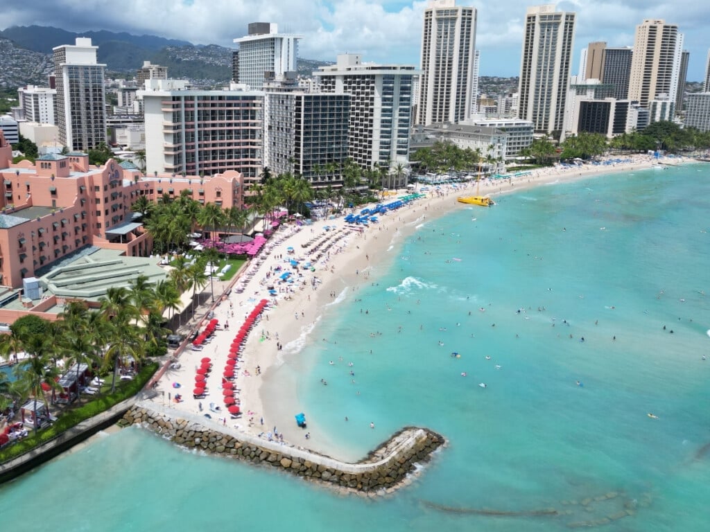 Aerial view of Royal Moana Beach (also called Royal Hawaiian Beach) in Waikīkī with vibrant blue ocean. You can see the pink Royal Hawaiian Hotel building and red and blue colored umbrellas line the sand.