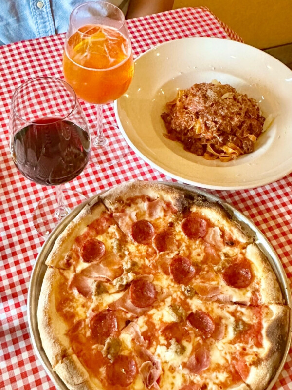 A pizza and pasta with Aperol Spritz and red wine on a red and white tablecloth taken at Arancino on Beachwalk in Waikīkī.