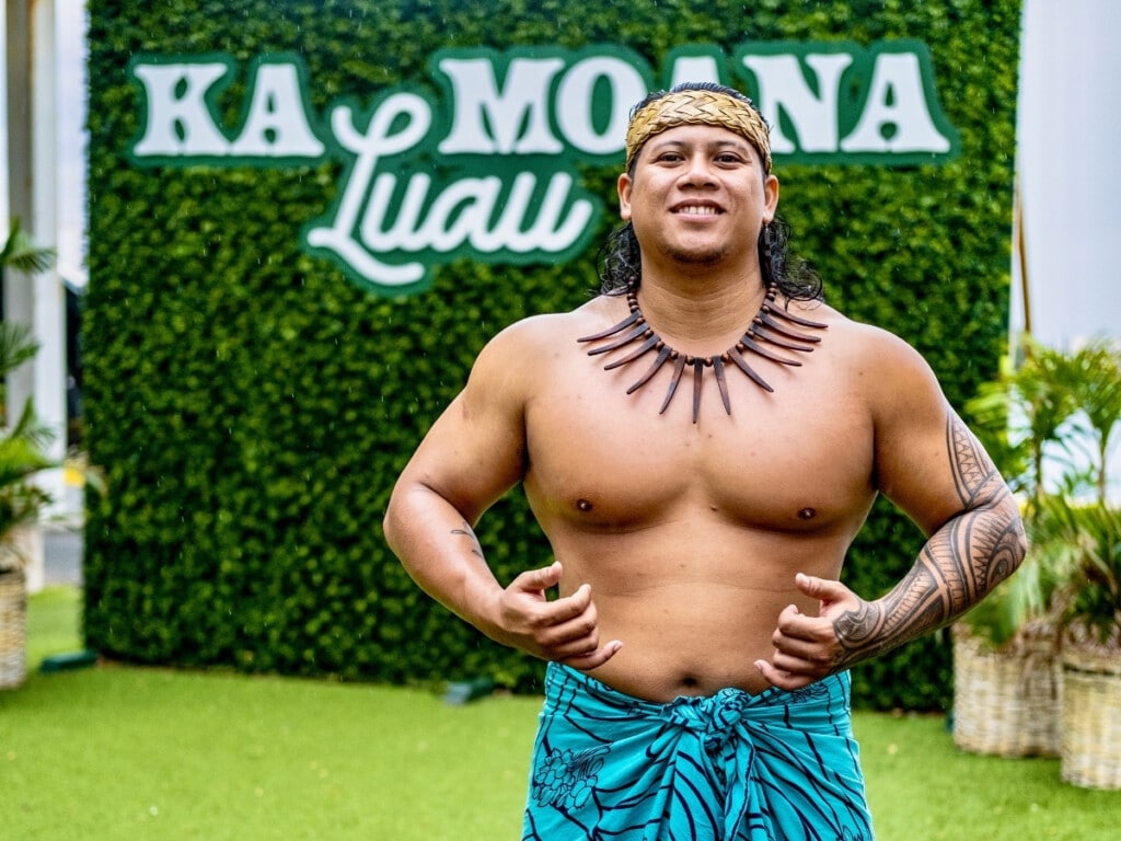 A performer in vibrant Polynesian attire strikes a pose in front of the iconic Ka Moana backdrop.