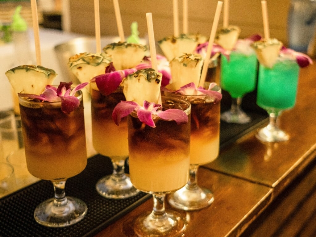 Generously sized Mai Tais and vibrant Blue Hawaiis sit pretty at the bar at Nā Lei Aloha lūʻau, garnished with fresh pineapple slices and orchids.