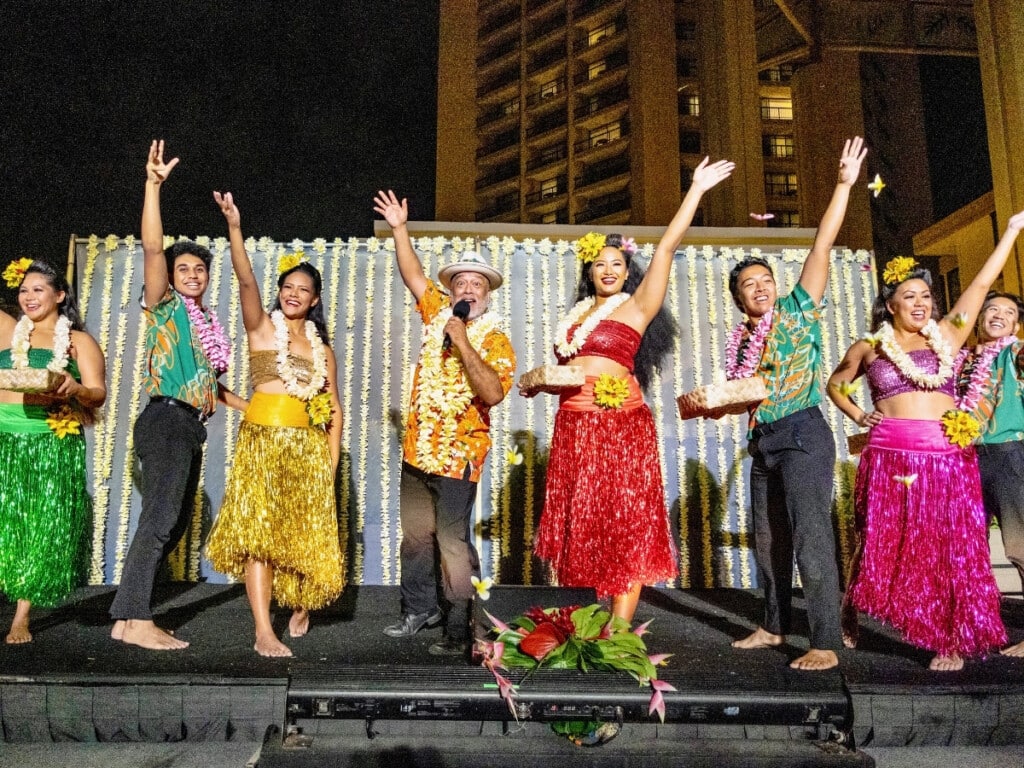 The performers on stage waving to the crowd with the MC in the middle at the Nā Lei Aloha Waikīkī lūʻau.